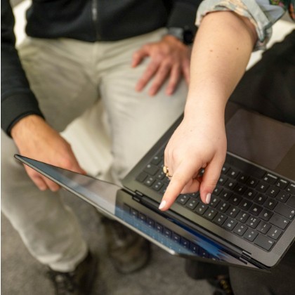 A close view of a right hand pointing to the screen of a laptop on a person’s lap. Another person sits next to them facing in the direction of the laptop. The scene represents the sharing of information.