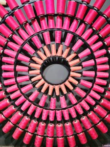 A striking circular arrangement of bright pink and orange thread spools creates a strong geometric pattern. The repeated shapes form concentric rings around the center, producing an eye-catching abstract composition. Captured at Chhatrapati Shivaji Maharaj Vastu Sangrahalaya, Mumbai, Maharashtra.