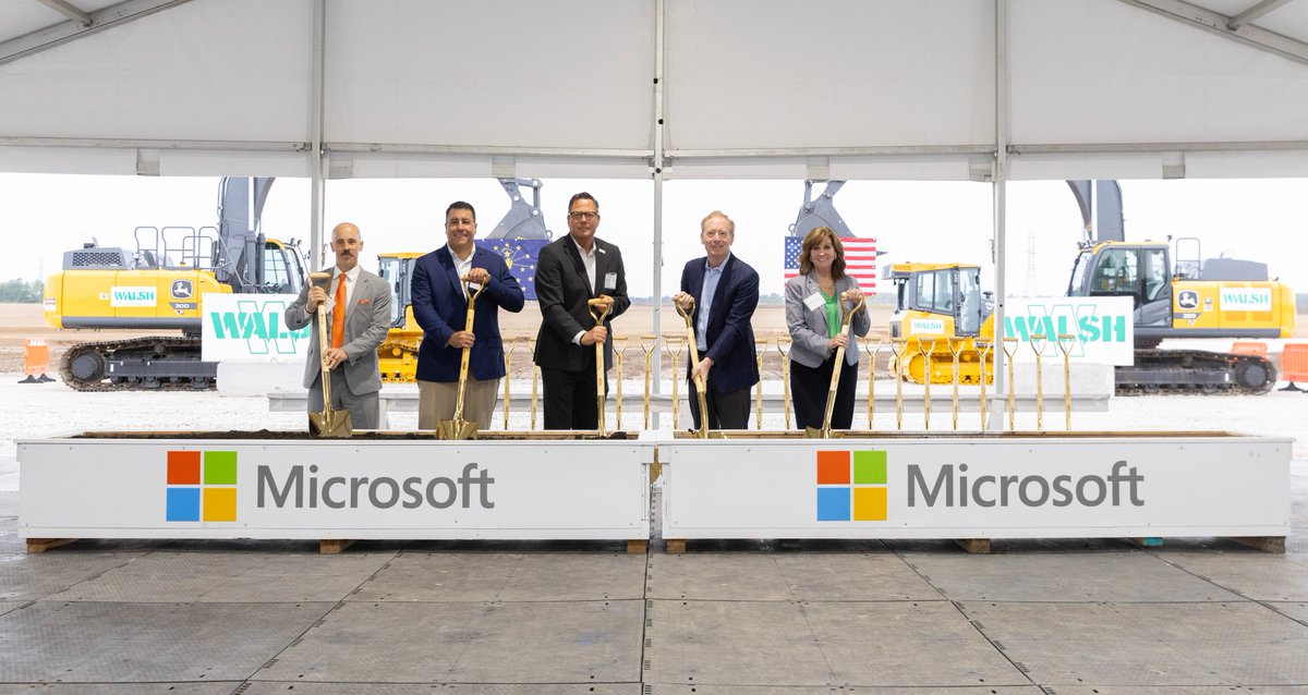 People stand in a row inside a large construction tent, each holding a ceremonial shovel at a groundbreaking event. Behind them are construction vehicles and signs, and in front are barriers displaying the Microsoft logo.