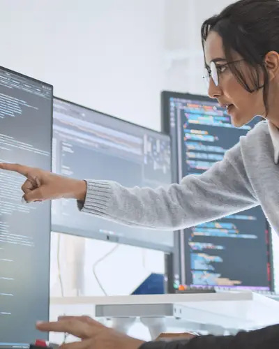 Person pointing at code on monitor in a bright office.
