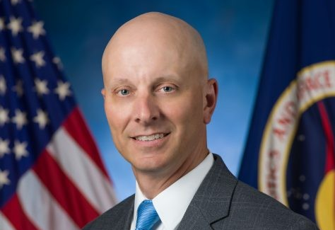 Steve Koerner, a white man, in a dark suit and blue tie, stands before the American and NASA flags