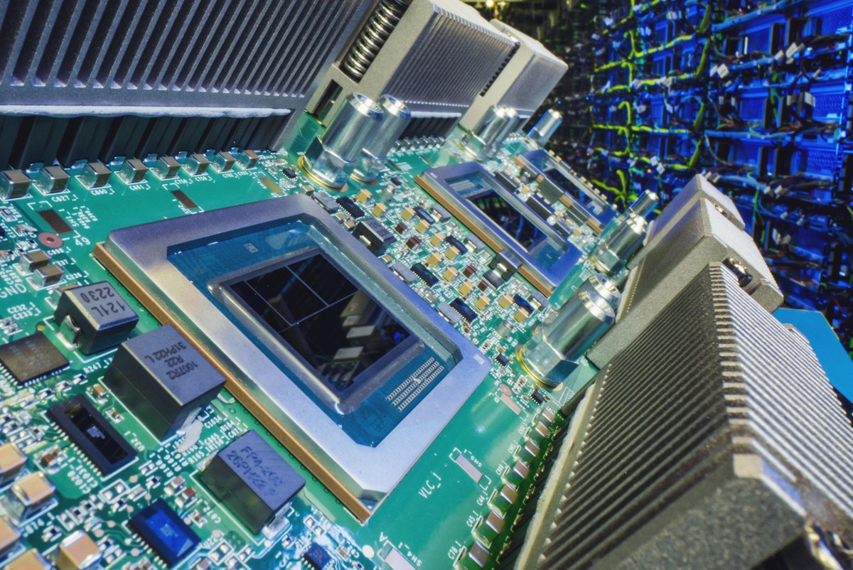 A close-up, angled view shows a green printed circuit board featuring large square processors, various electronic components, and silver heat sinks. Text on components includes "121L 2230" and "FP4-200," with labels like "VLC_1" printed on the board. The background displays an out-of-focus data center with many server racks illuminated by blue and green lights.